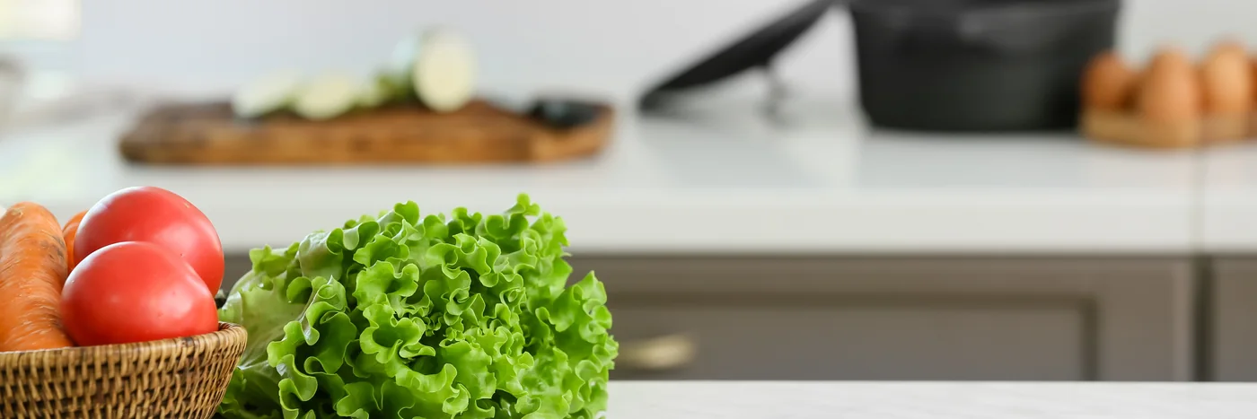 A basket of tomatoes and lettuce on a bright kitchen counter, with vegetables and cookware behind.